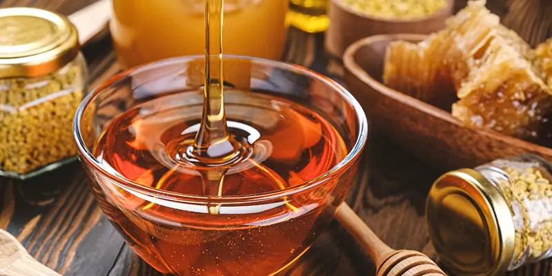 Honig tropft aus Holzlöffel in Glasschale, umgeben von Bienenwaben und anderen Heilmitteln